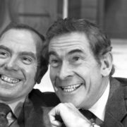 Two men in suits smiling and posing closely together for the camera in a black and white photograph.