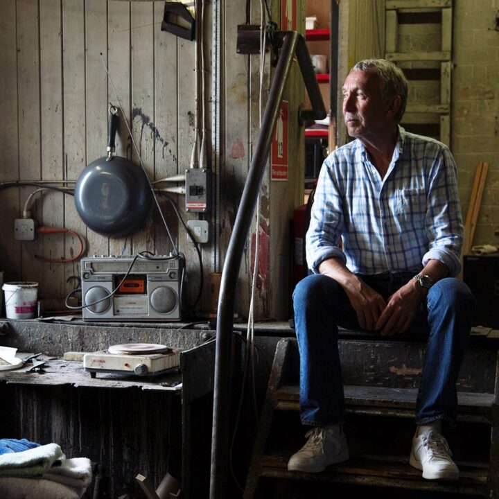 A man in a plaid shirt and jeans sits on steps inside a cluttered workshop, with tools, equipment, and a radio visible around him.