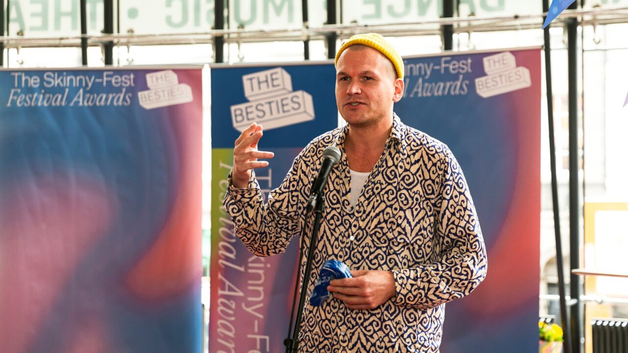 A man in a patterned shirt and yellow beanie speaks into a microphone at The Skinny-Fest Festival Awards, standing in front of event banners.