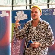 A man in a patterned shirt and yellow beanie speaks into a microphone at The Skinny-Fest Festival Awards, standing in front of event banners.