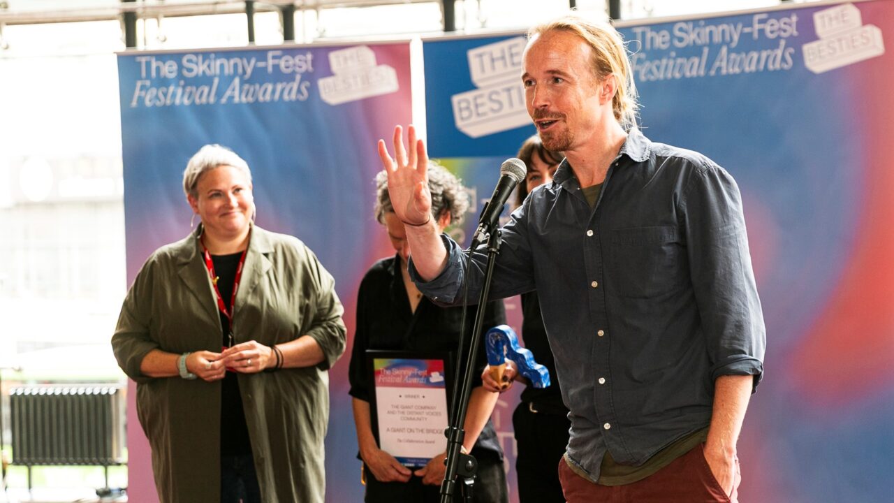 A man speaks into a microphone at an awards event while others stand behind him, with "The Skinny-Fest Festival Awards" banners in the background.