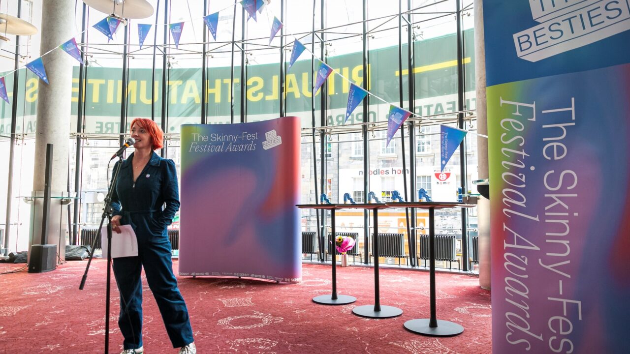 A person with short red hair speaks into a microphone at The Skinny-Fest Festival Awards, standing in front of event banners and round tables in a modern venue.