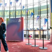 A person with short red hair speaks into a microphone at The Skinny-Fest Festival Awards, standing in front of event banners and round tables in a modern venue.