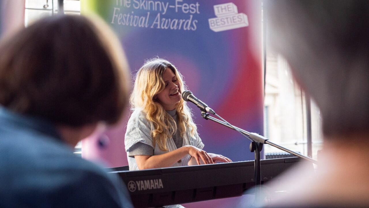 A woman plays a Yamaha keyboard and sings into a microphone at an indoor event with a "Festival Awards" banner in the background.