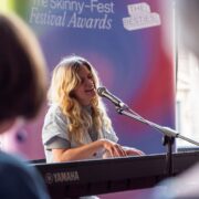 A woman plays a Yamaha keyboard and sings into a microphone at an indoor event with a "Festival Awards" banner in the background.