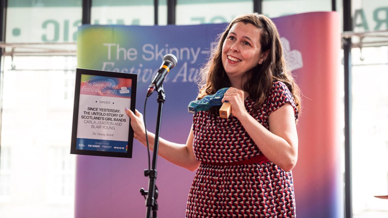 A woman stands on stage holding a framed award and a blue object, smiling in front of a microphone and a backdrop with "The Skinny" festival logo.