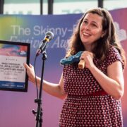 A woman stands on stage holding a framed award and a blue object, smiling in front of a microphone and a backdrop with "The Skinny" festival logo.