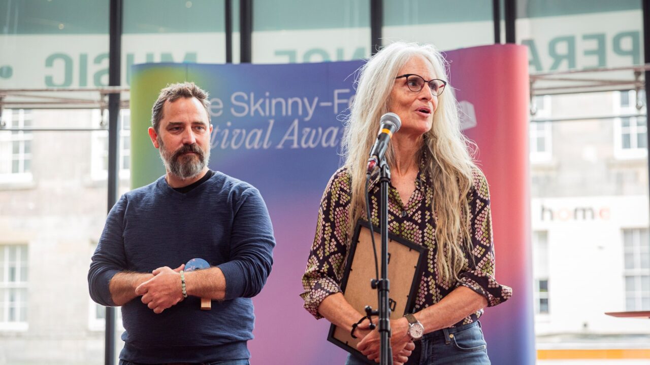 A man and a woman stand on stage; the woman speaks at a microphone holding a plaque, while the man stands beside her holding an object. A colourful event backdrop is behind them.