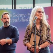 A man and a woman stand on stage; the woman speaks at a microphone holding a plaque, while the man stands beside her holding an object. A colourful event backdrop is behind them.
