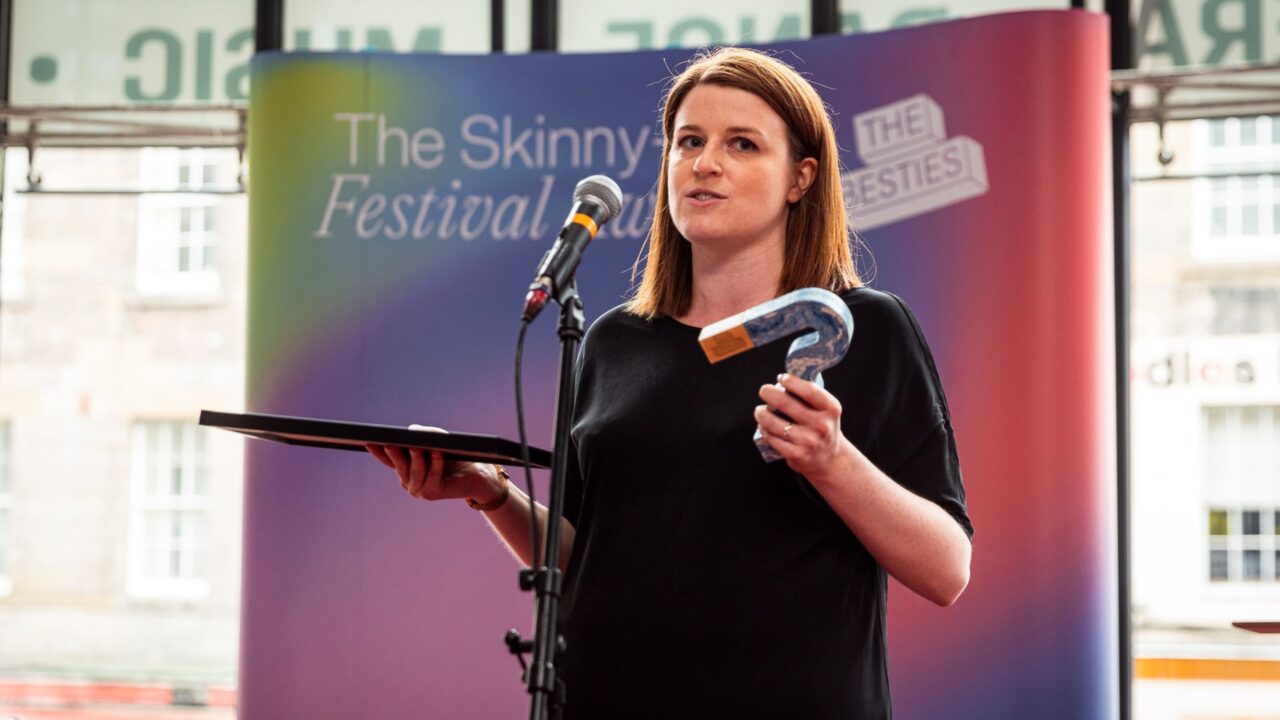 A woman stands on stage holding a framed certificate and a question mark-shaped award, speaking into a microphone at The Skinny Festival event.