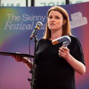 A woman stands on stage holding a framed certificate and a question mark-shaped award, speaking into a microphone at The Skinny Festival event.