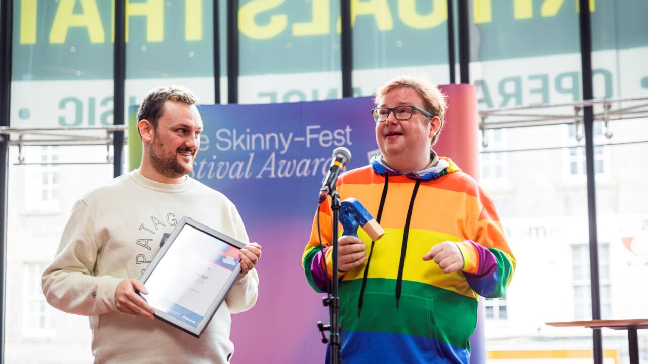 Two people stand indoors at an awards event; one holds a framed certificate, the other holds a trophy and speaks into a microphone, wearing a rainbow-striped hoodie.