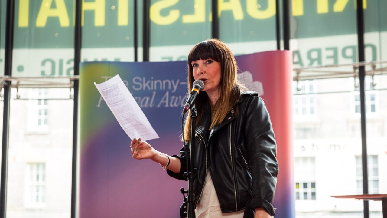 A woman in a black leather jacket speaks into a microphone and holds a sheet of paper on a stage with a colourful backdrop.