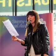 A woman in a black leather jacket speaks into a microphone and holds a sheet of paper on a stage with a colourful backdrop.