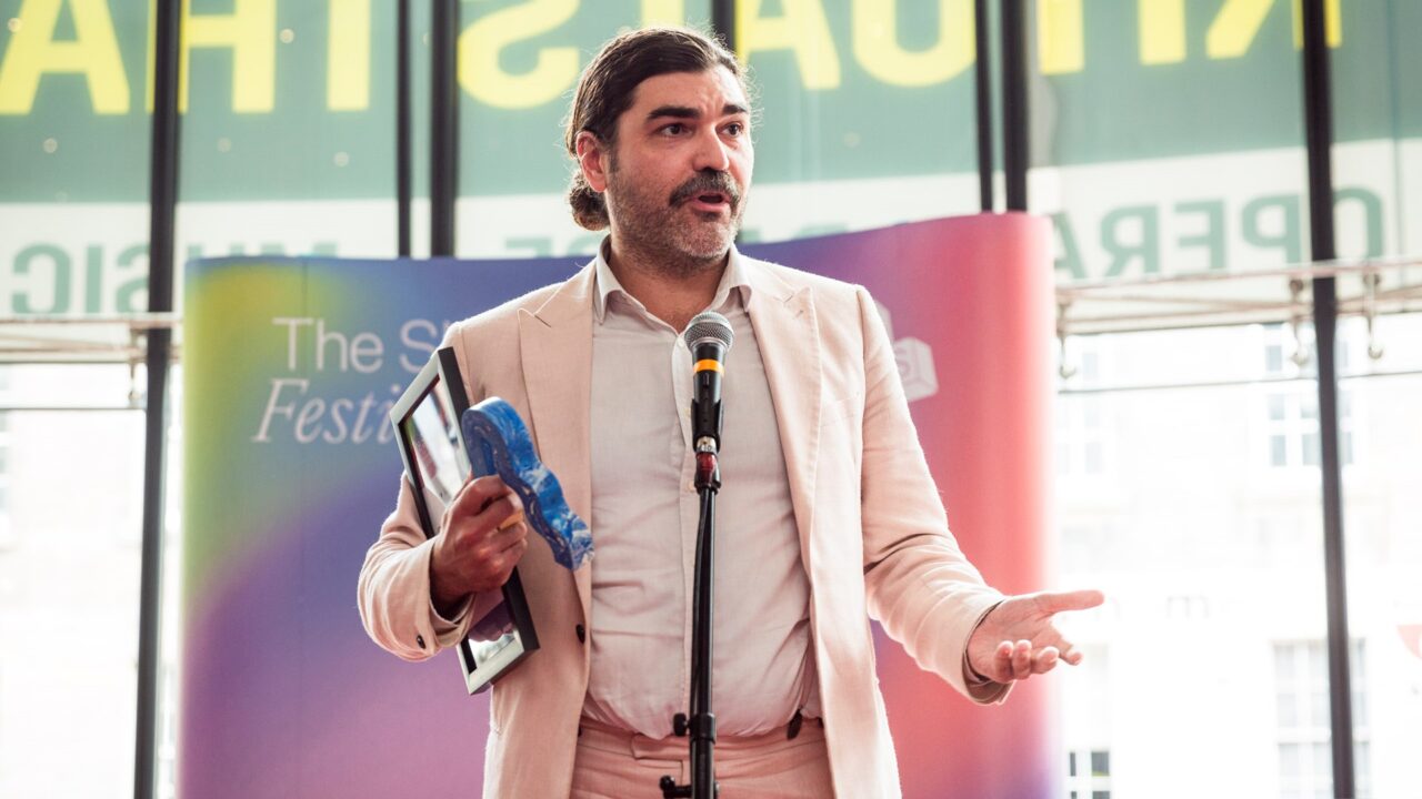 A man in a light pink suit stands at a microphone holding a framed certificate and a blue object, speaking at an indoor event with a colourful backdrop.