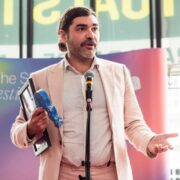 A man in a light pink suit stands at a microphone holding a framed certificate and a blue object, speaking at an indoor event with a colourful backdrop.