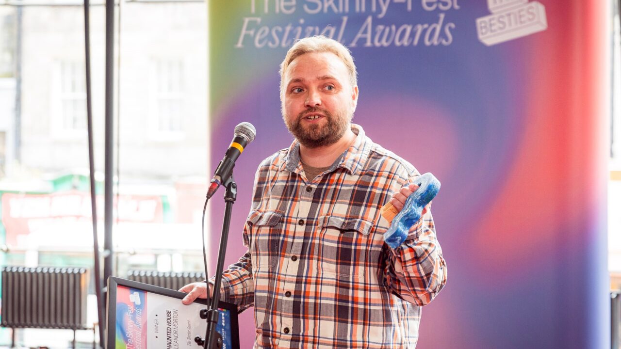 A man in a plaid shirt stands at a microphone holding an award and a framed certificate at The Skinny Fest Festival Awards.