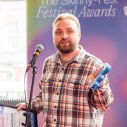 A man in a plaid shirt stands at a microphone holding an award and a framed certificate at The Skinny Fest Festival Awards.
