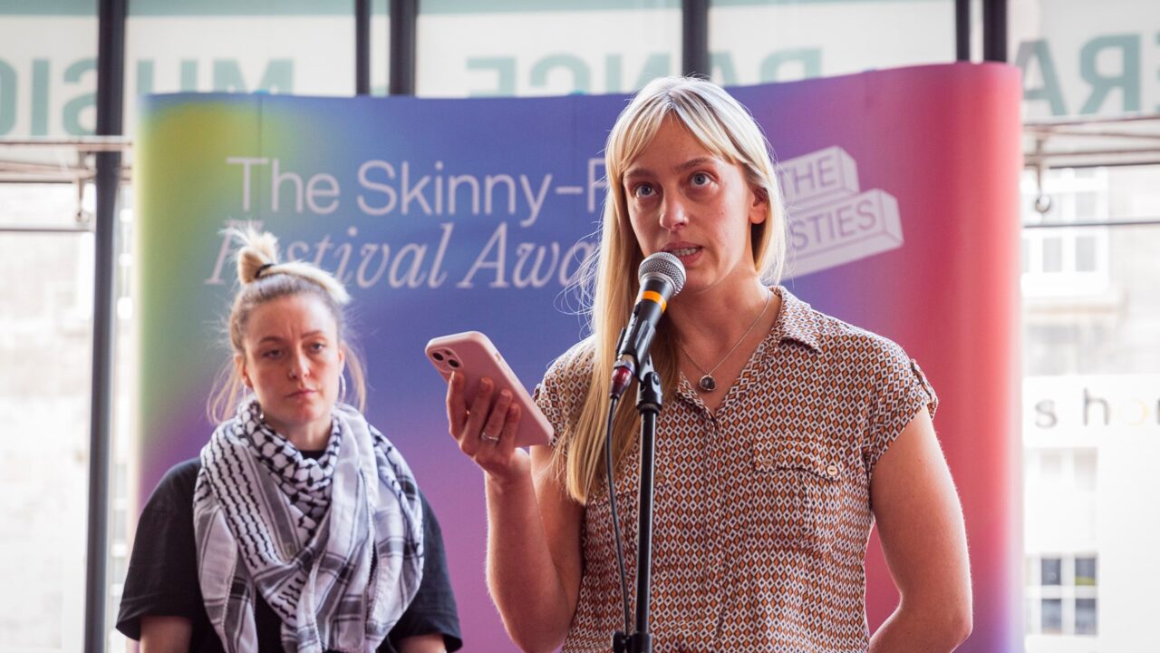 Two women stand at microphones in front of a colourful backdrop that reads "The Skinny Festival Awards." One woman is speaking while holding a phone.