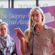 Two women stand at microphones in front of a colourful backdrop that reads "The Skinny Festival Awards." One woman is speaking while holding a phone.