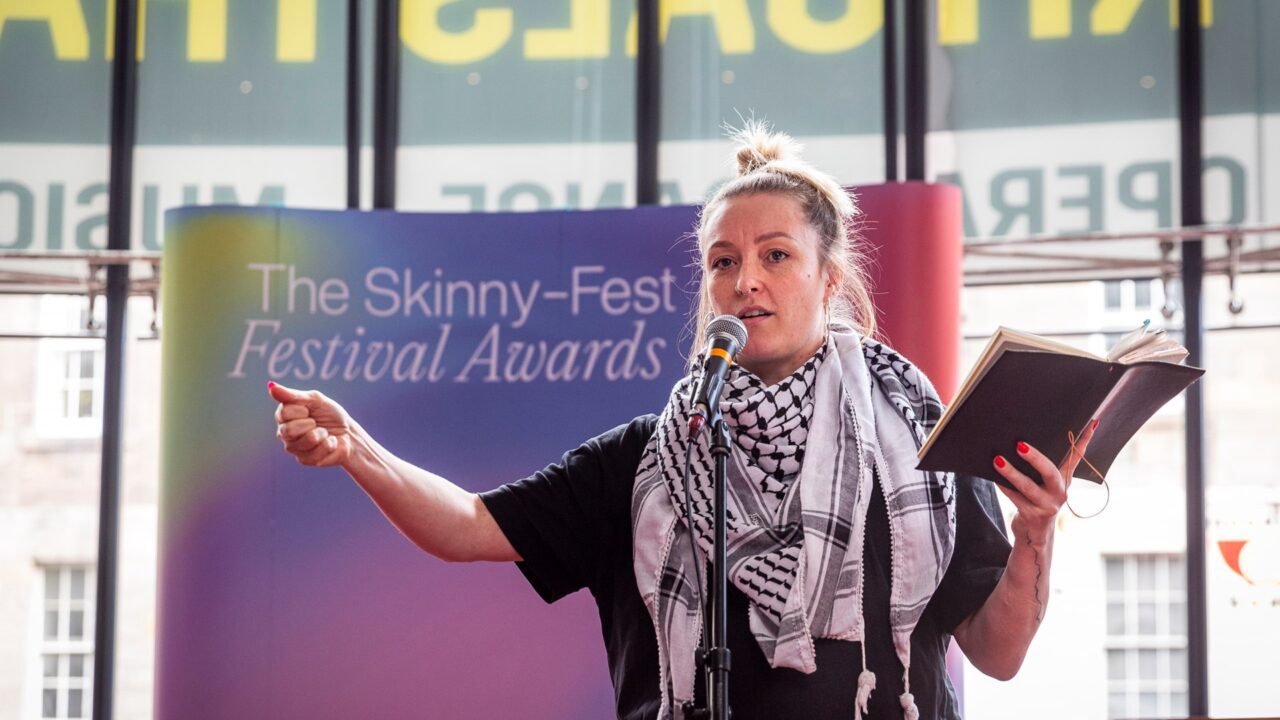 A person speaks into a microphone while holding a notebook on stage at The Skinny Fest Festival Awards event.