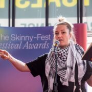 A person speaks into a microphone while holding a notebook on stage at The Skinny Fest Festival Awards event.