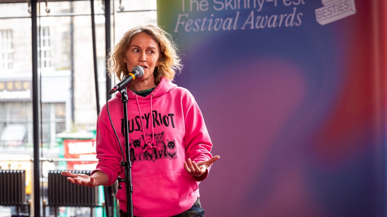 A person in a pink "Pussy Riot" hoodie speaks into a microphone on stage at The Skinny Fest Festival Awards event.