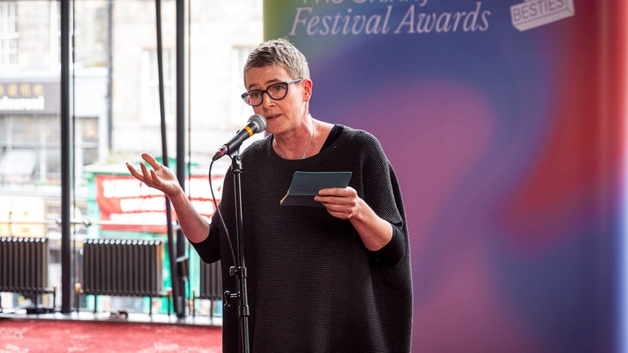 A person with short grey hair and glasses speaks into a microphone while holding notes at an event labelled "Festival Awards.