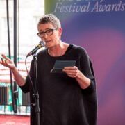 A person with short grey hair and glasses speaks into a microphone while holding notes at an event labelled "Festival Awards.