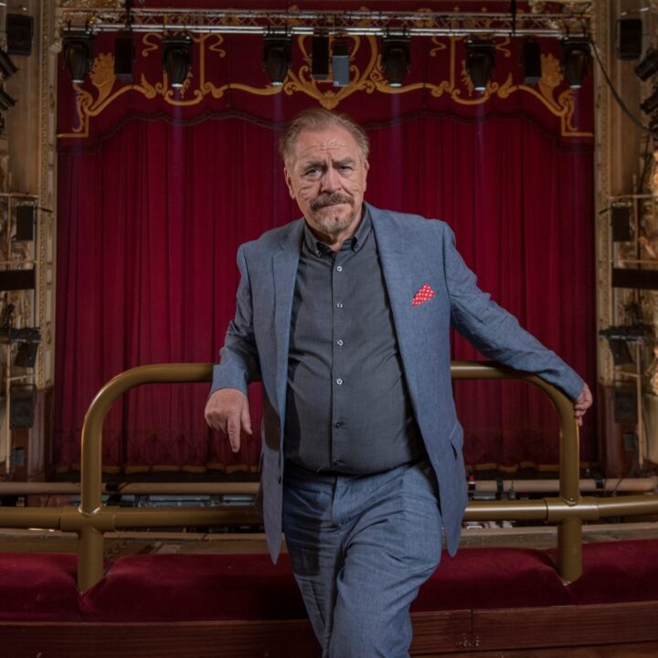 A man in a blue suit stands in a theatre balcony with red velvet seats, ornate lighting, and a large red curtain on stage in the background.
