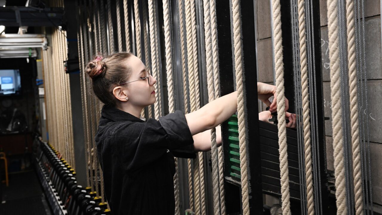A person in black clothing adjusts stage rigging ropes backstage in a theater, surrounded by multiple vertical ropes and weights.