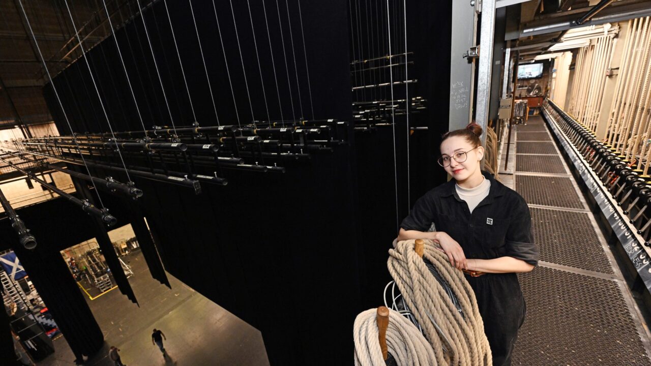 A stagehand stands on a backstage beside rigging ropes and stage equipment, overlooking the theatre below.