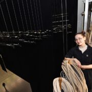 A stagehand stands on a backstage beside rigging ropes and stage equipment, overlooking the theatre below.