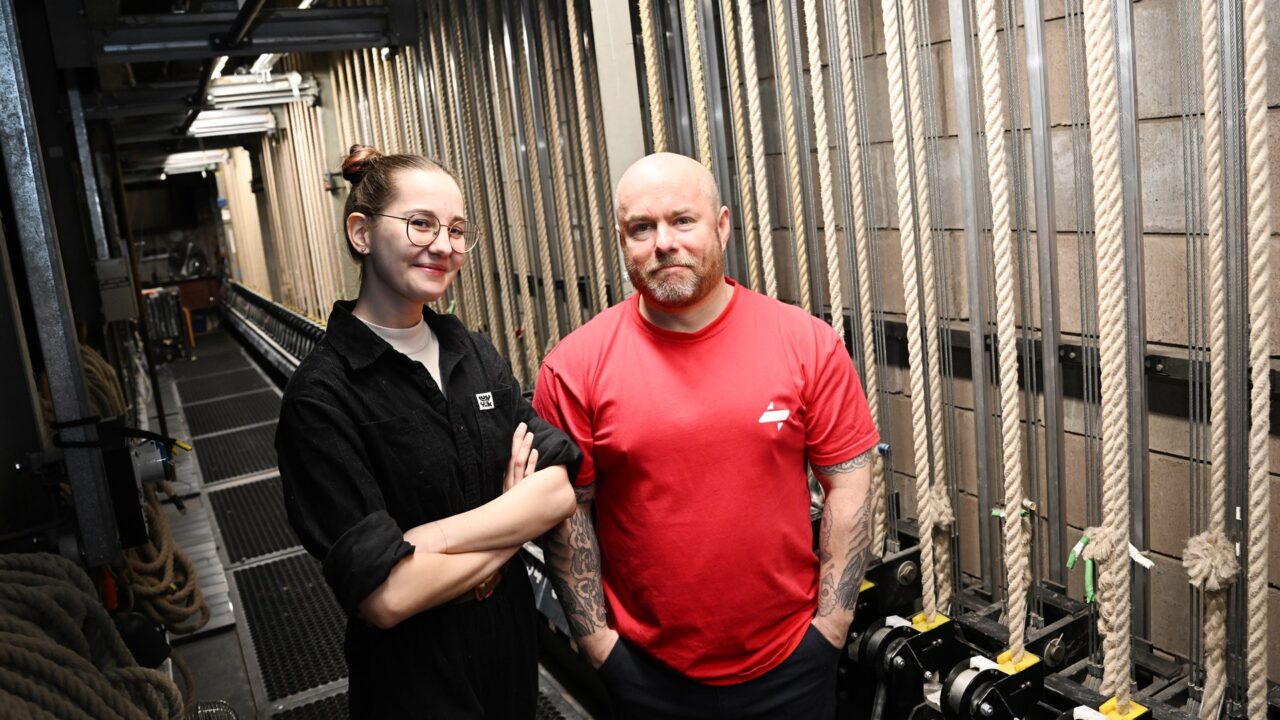 Two people stand side by side in a backstage area with ropes and stage equipment, facing the camera.