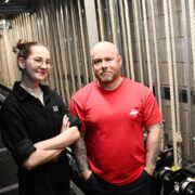 Two people stand side by side in a backstage area with ropes and stage equipment, facing the camera.