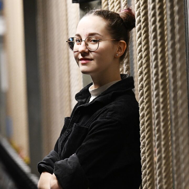 A person with glasses and hair in a bun leans against a wall with thick ropes, arms crossed, looking at the camera.
