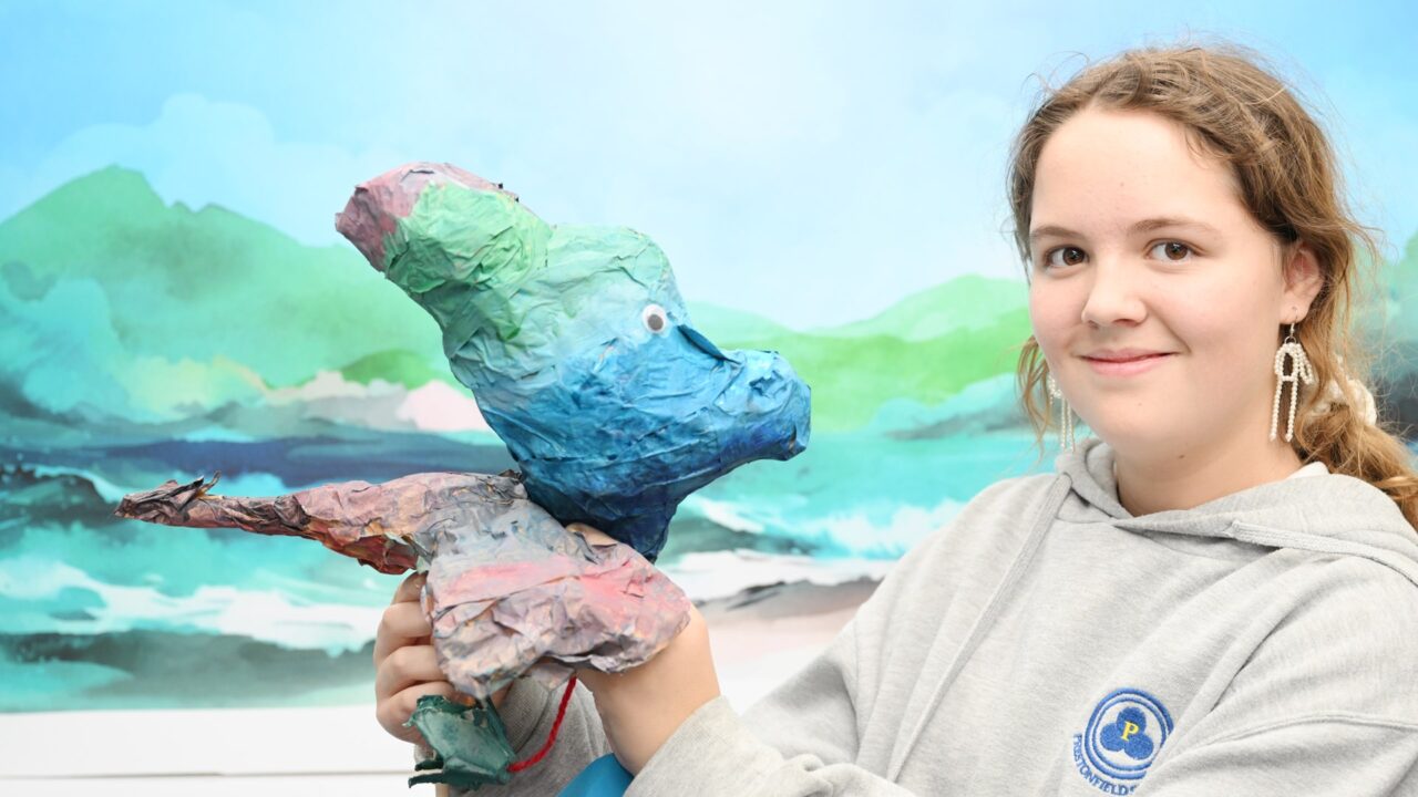 A young person in a grey hoodie holds a colorful papier-mâché animal sculpture, with a painted landscape backdrop behind them.