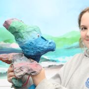 A young person in a grey hoodie holds a colorful papier-mâché animal sculpture, with a painted landscape backdrop behind them.