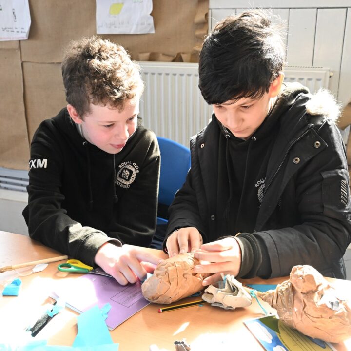 Two boys sit at a table in a classroom, working together on a papier-mâché model with craft materials scattered around them.