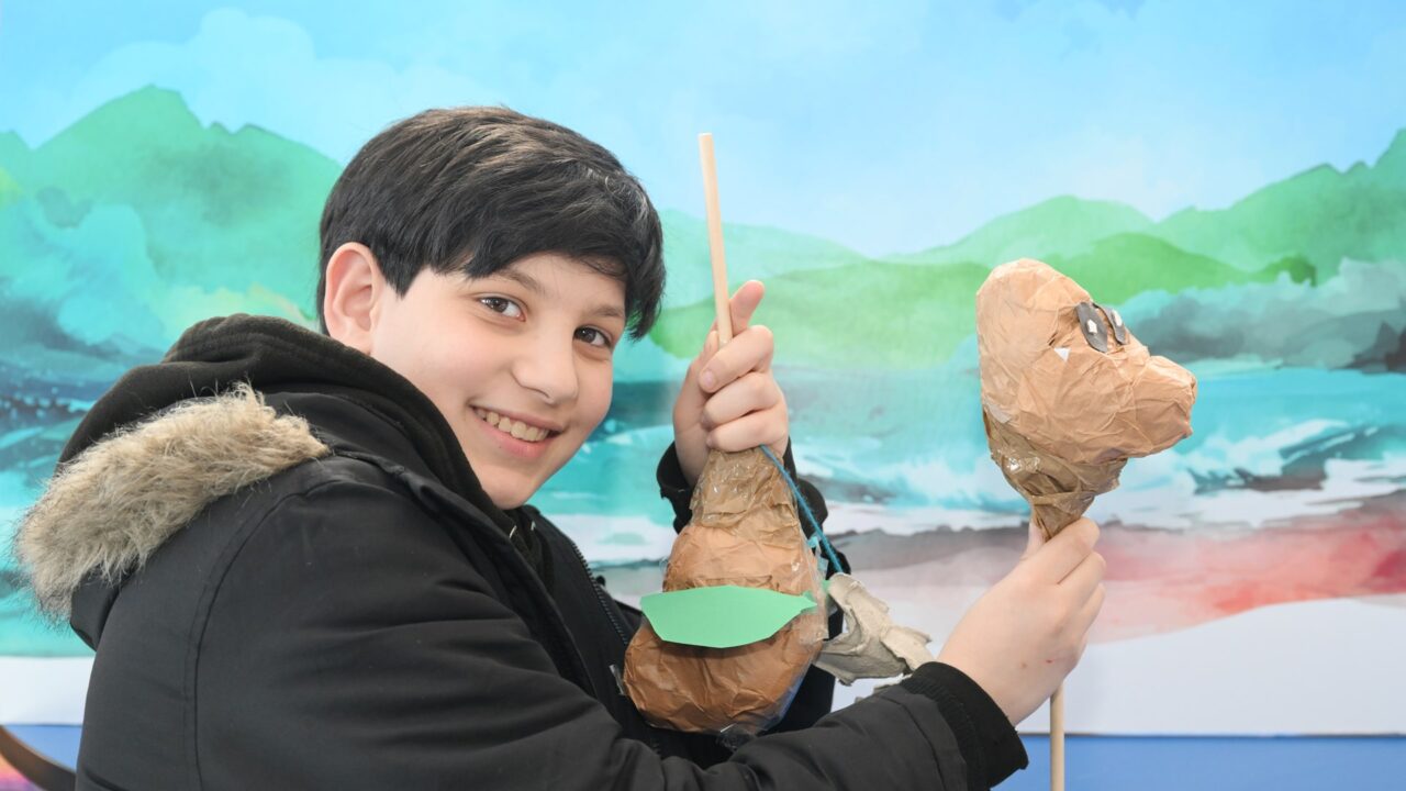 A smiling child in a black coat holds up two paper mache puppets in front of a painted backdrop of mountains and water.