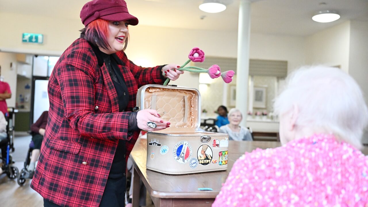 A performer in a red plaid jacket and hat presents a fabric flower from a suitcase to an older person seated at a table in a communal room.