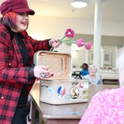 A performer in a red plaid jacket and hat presents a fabric flower from a suitcase to an older person seated at a table in a communal room.