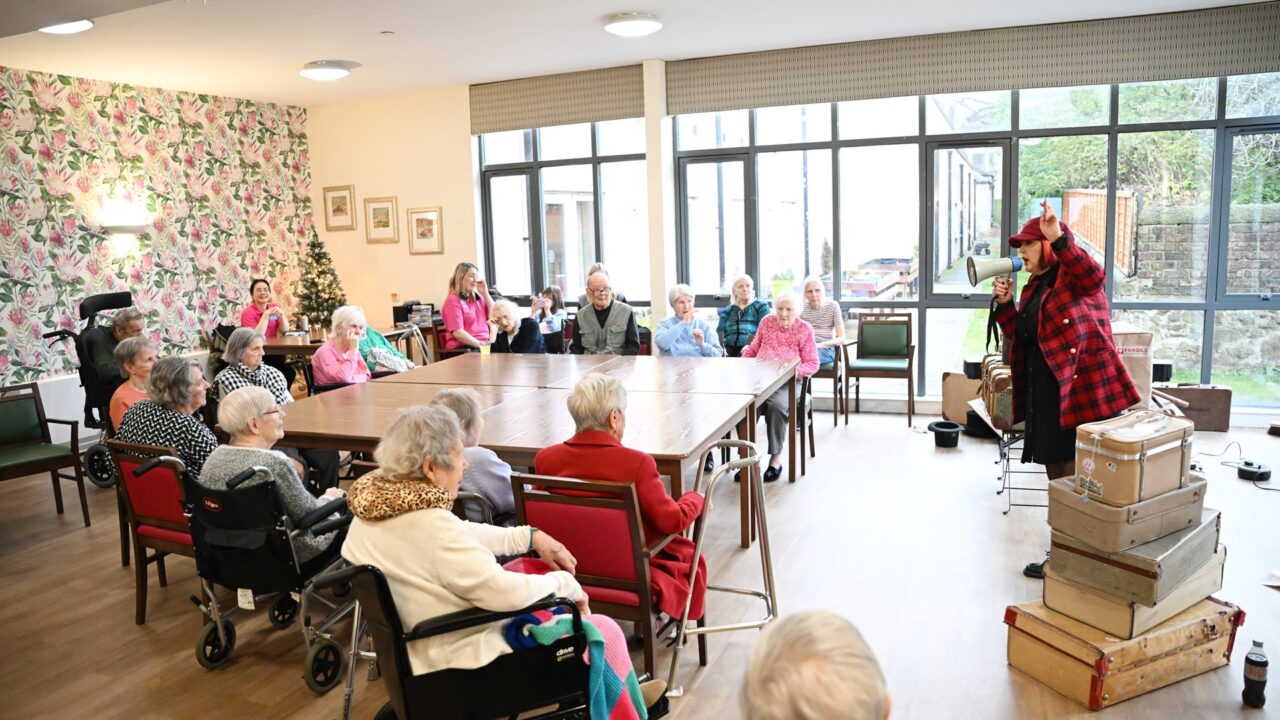 A group of elderly people sit in a common room watching a performer in a red plaid shirt and hat, with stacked suitcases nearby.