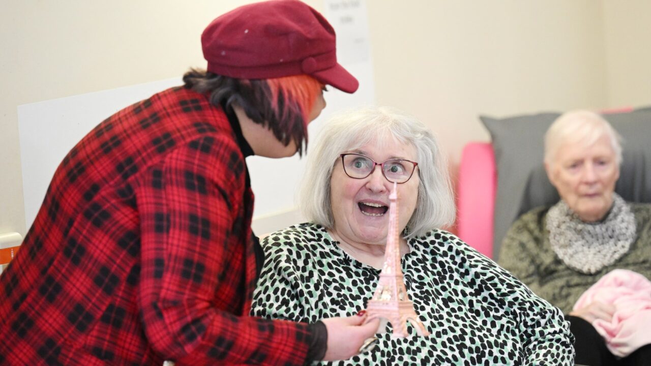 A person shows a small Eiffel Tower model to an elderly woman who looks surprised and happy; another elderly woman sits in the background.