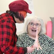 A person shows a small Eiffel Tower model to an elderly woman who looks surprised and happy; another elderly woman sits in the background.