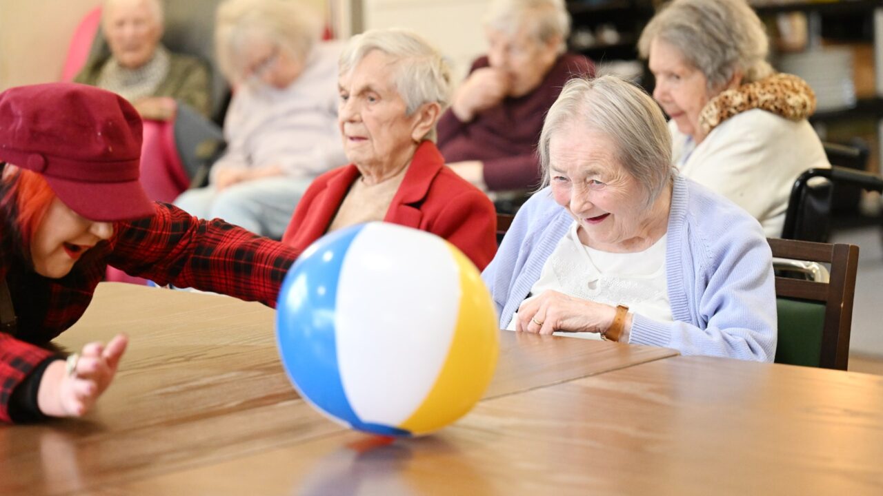 Elderly women sitting around a table playing with a colourful beach ball in a communal setting.