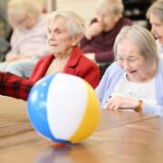 Elderly women sitting around a table playing with a colourful beach ball in a communal setting.