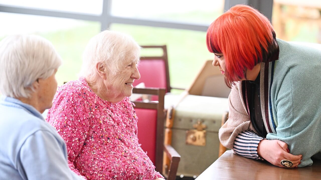 A woman with red hair leans on a table, talking to two elderly women sitting next to each other, one wearing a pink sweater, indoors.