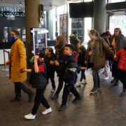 A group of adults and children, some holding hands, walk through a lobby area with large glass windows and columns.