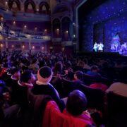 Audience members watch a theatrical performance on stage inside a large, ornate theatre with balconies and decorative lighting.